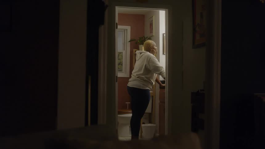 A woman in a white hoodie stands at a brightly lit bathroom sink framed by a dark hallway at night