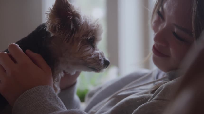 A woman in a hoodie smiles softly at a small dog she holds close near a window