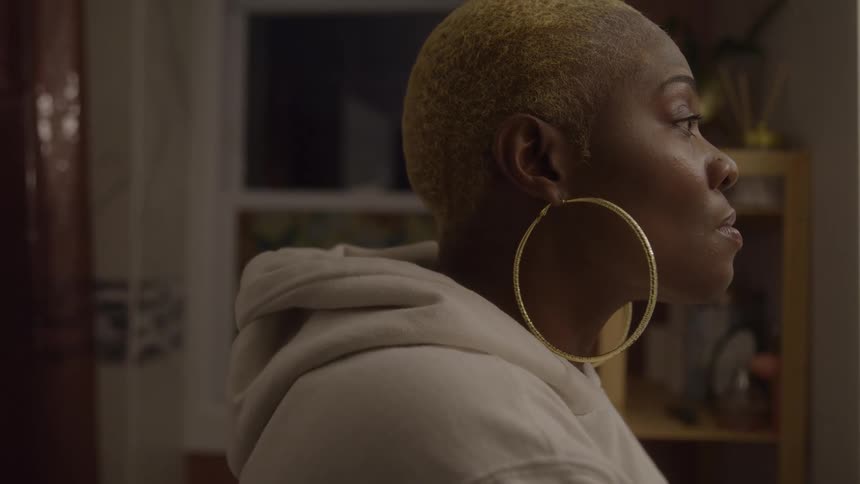 A woman with very short blonde hair and large hoop earrings in a hoodie gazes sadly and thoughtfully