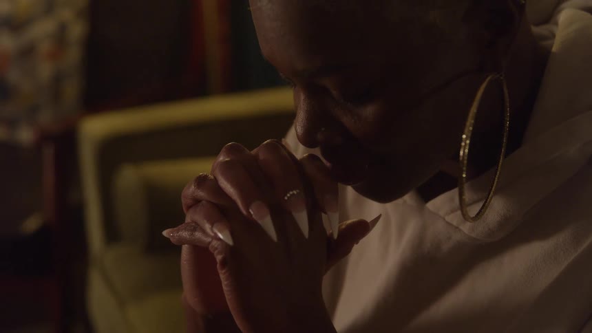 A dramatic dark close view shows a woman with short hair wearing a white hoodie and large gold hoop earrings sitting with her hands clasped and long acrylic nails looking down in a solemn mood