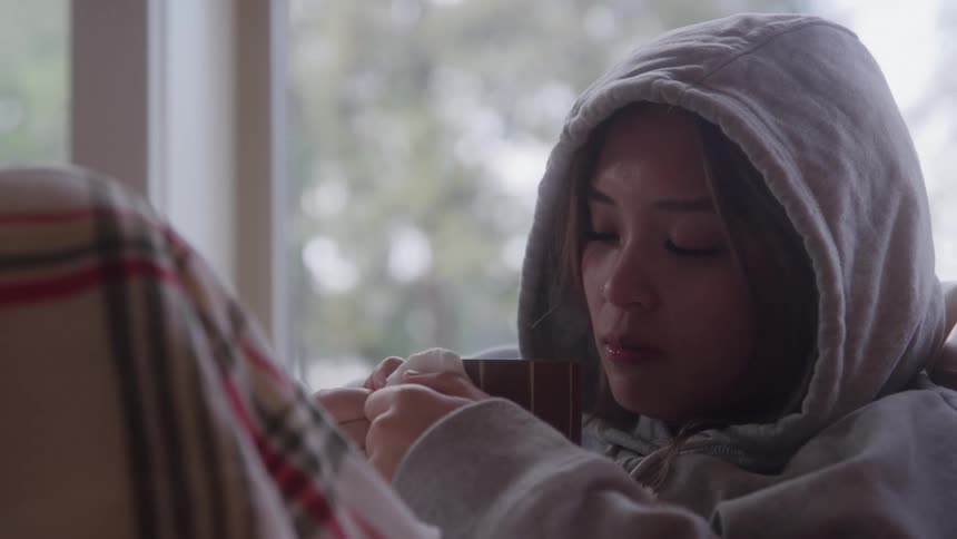 A sick woman sips hot coffee wrapped in a blanket indoors