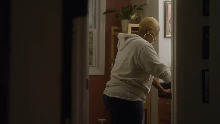 A woman with short blonde hair wearing a white hoodie and large hoop earrings stands bent over a sink viewed from a dark hallway looking into a brightly lit bathroom with a window and houseplants