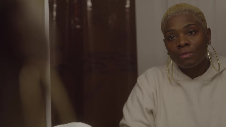 A young woman stands in front of a mirror her short hair sprayed gold and adorned with large round golden earrings gazing at her reflection with a sad expression