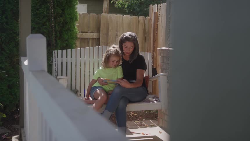 Young mother and daughter reading together on a swing outdoors on the lawn