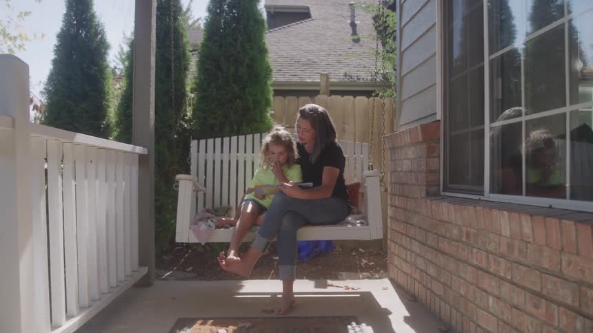 Mother and daughter read on a porch swing outside a home at midday