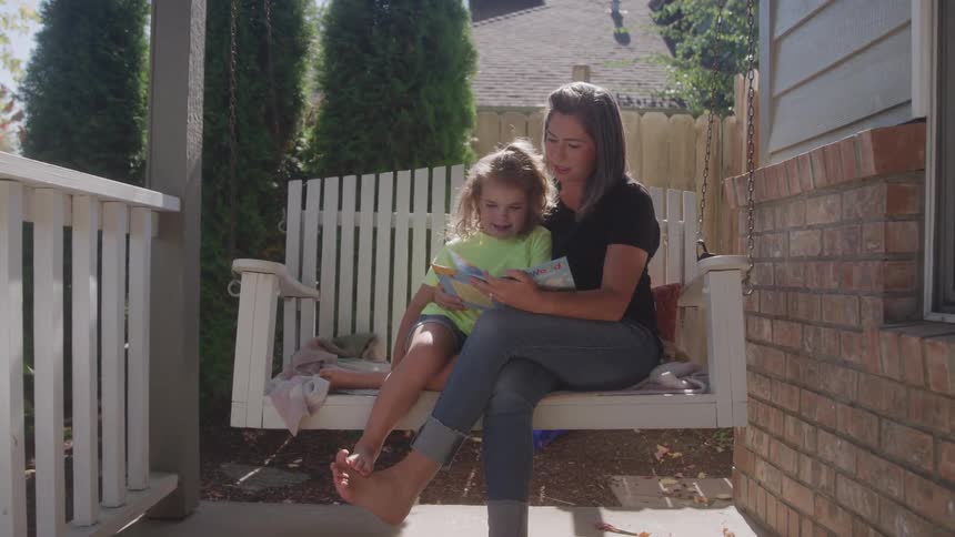 A mother and daughter sit together on an outdoor swinging chair beside a house at midday reading a storybook under the warm sun