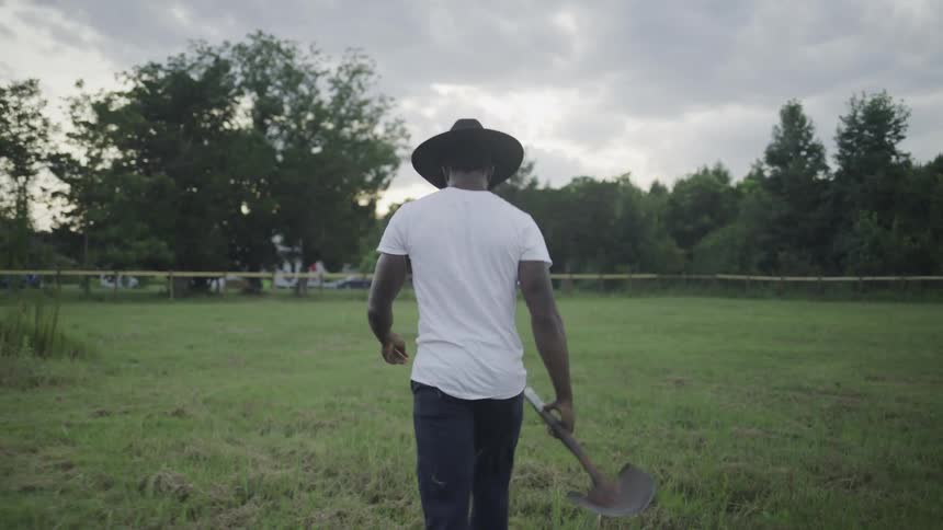 The video shows a man from behind wearing a white tshirt and a dark cowboy hat standing in a grassy field surrounded by trees he is holding his hat in his right hand and has a serious or contemplative posture as he looks out into the landscape