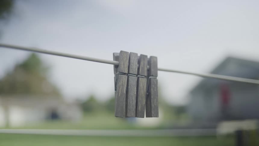 The video is a shallow focus closeup of three wooden clothespins clipped onto a taut clothesline the background is blurred showing a house and a yard under an overcast sky