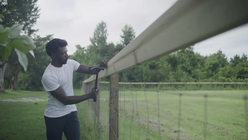 The video is a medium shot of a man in a white tshirt and dark pants using a hammer to fix a wire fence on a bright overcast day the scene is set outdoors with a line of trees and green foliage in the background