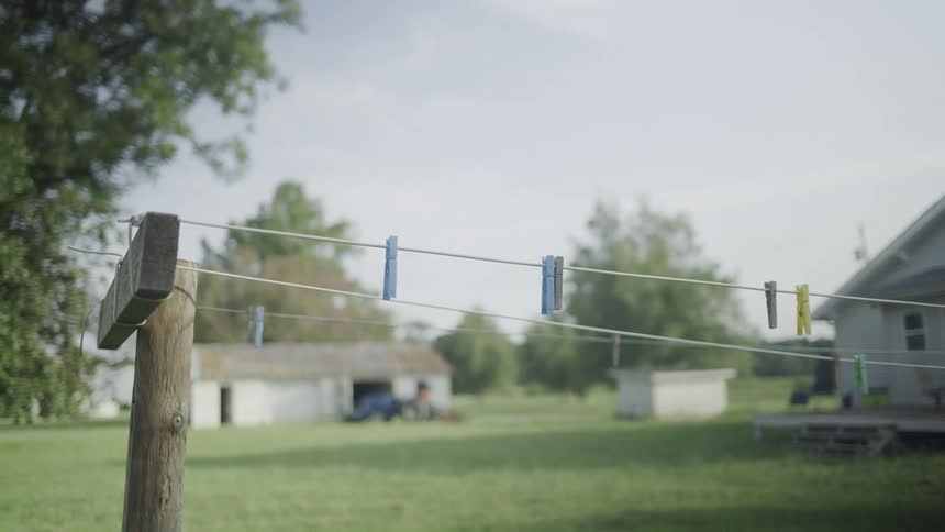 A quiet yard shows three empty clotheslines with colorful clothespins clipped along them as small outbuildings sit in the soft distance