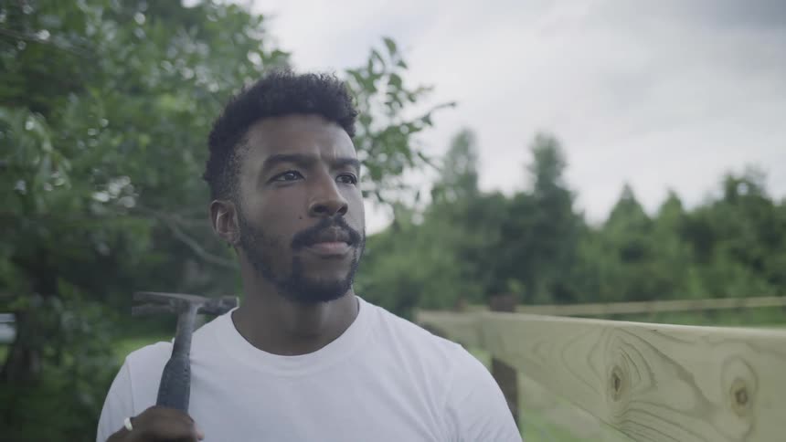 A closeup of a man wearing a white shirt shows his profile as he stands outdoors holding a hammer over his shoulder and looking off into the distance