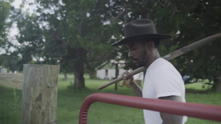 The video is a mediumclose shot of a man wearing a white tshirt and a dark cowboy hat he is carrying a shovel over his shoulder as he walks outdoors past a wooden fence toward a house in the background