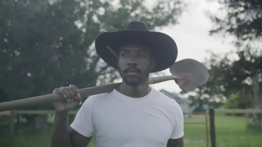 A young man walks across a sunlit lawn at midday wearing a sunhat and carrying a shovel over his shoulder