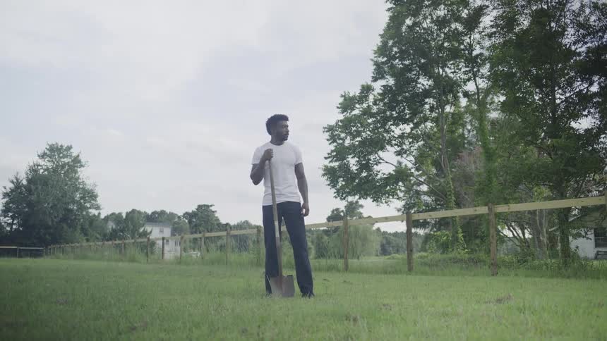The video is a mediumlong shot of a man wearing a white tshirt and dark pants standing in a grassy field on a cloudy day he is holding a shovel which rests on the ground beside him with a wooden fence line and scattered trees in the background