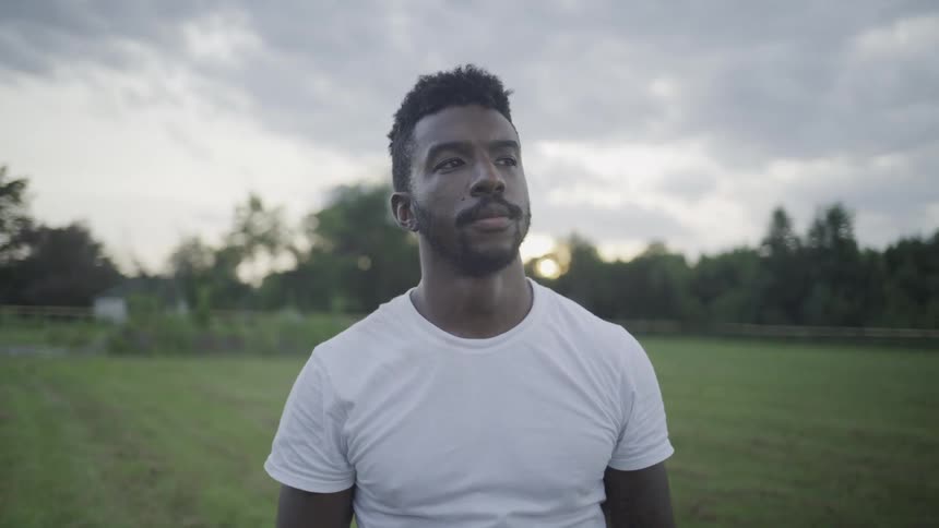 The video shows a man outdoors wearing a white tshirt who removes a dark cowboy hat and runs his hand over his forehead as if wiping sweat he is standing in a grassy field with trees and a cloudy sky in the background