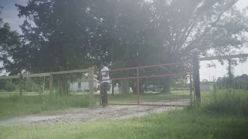 The video is a wide shot of a man in a white tshirt and dark pants walking toward a red metal gate on a dirt path he is opening the gate to an enclosed area with a large tree and a house in the background
