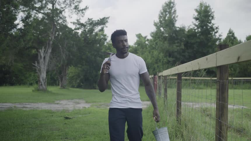 A man wearing a white shortsleeved shirt and dark trousers walks along a line of wooden posts and wire fencing on a grassy field carrying a metal hammer on his shoulder and a small bucket in his hand