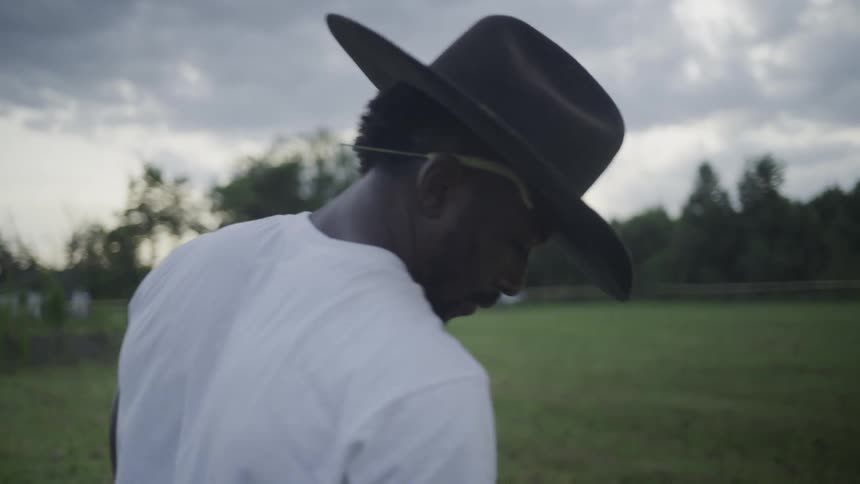 The video is a mediumclose shot of a man wearing a white tshirt and a dark cowboy hat in an outdoor grassy field he is looking downward with the brim of his hat slightly obscuring his face