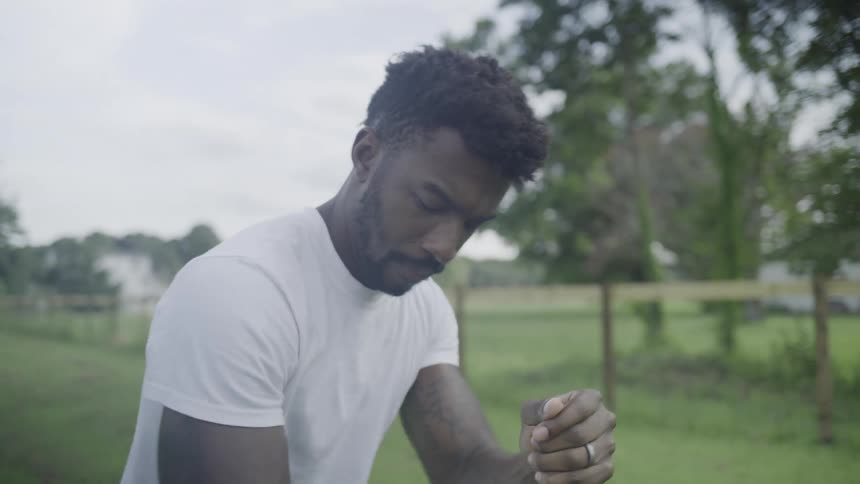 The video is a mediumclose shot of a man wearing a white tshirt and a dark cowboy hat he is carrying a shovel over his shoulder as he walks outdoors past a wooden fence toward a house in the background