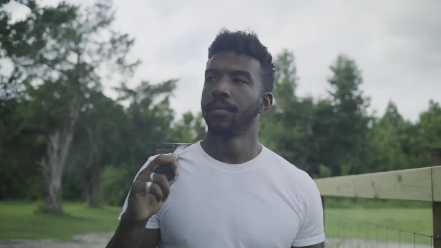 The video is a mediumclose shot of a man wearing a white tshirt standing outdoors with a small pair of clippers in his hand the man has a short dark haircut and a beard and is looking forward with a neutral expression against a backdrop o