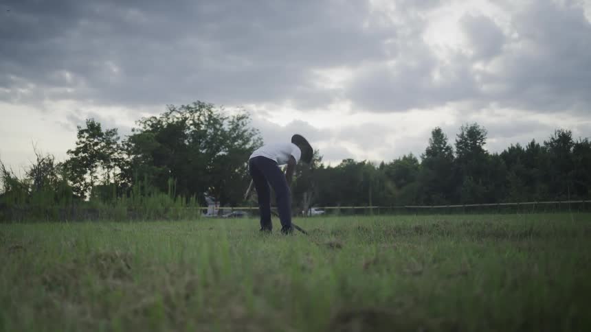 A man wearing a hat a white shirt and dark pants stands in the middle of a grassy field beneath a cloudy sky using a longhandled shovel to repeatedly dig into and turn over the earth