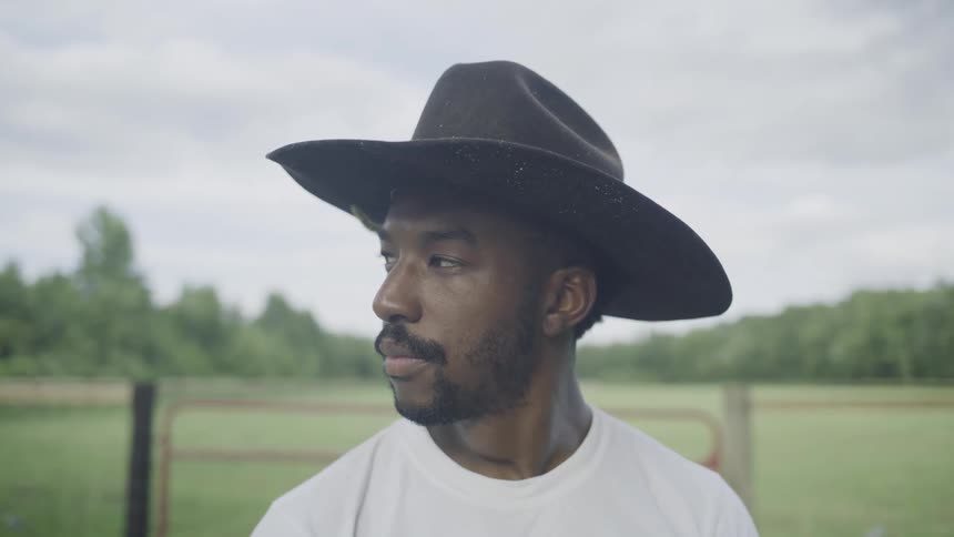 The video is a mediumclose shot of a man wearing a dark cowboy hat and a white tshirt standing outdoors he is looking to the side and then turns to look directly at the camera with a neutral serious expression