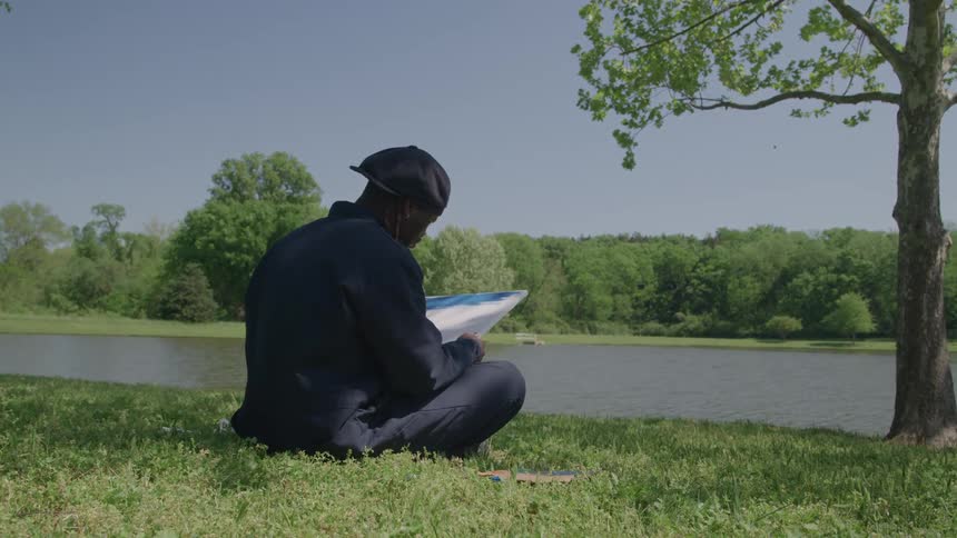 The video is a mediumlong shot of an african american man sitting on the grass near a pond or lake on a sunny day he is wearing a dark outfit and a black head covering while sketching or painting in a notebook resting on his lap