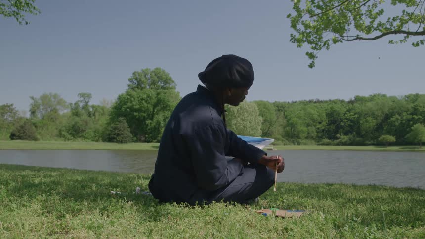 The video shows an african american man sitting in a grassy field next to a pond on a sunny day he is wearing a dark outfit and a black head covering while sketching or drawing in a notebook resting on his lap