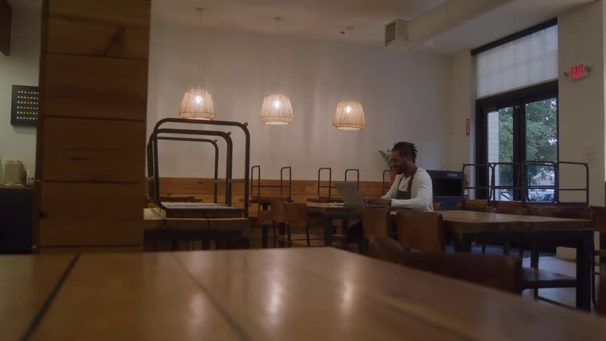 A man wearing an apron sits at a dark wooden table in a empty restaurant using a laptop computer while chairs are placed upside down on other nearby tables and three woven pendant lamps illuminate the area