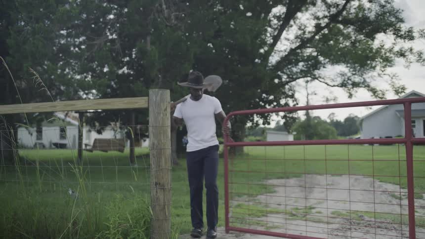 The video is a long shot of a man wearing a white tshirt and dark pants walking through a red metal gate and along a dirt driveway the scene is set on a farm or rural property with a house and large trees visible in the background