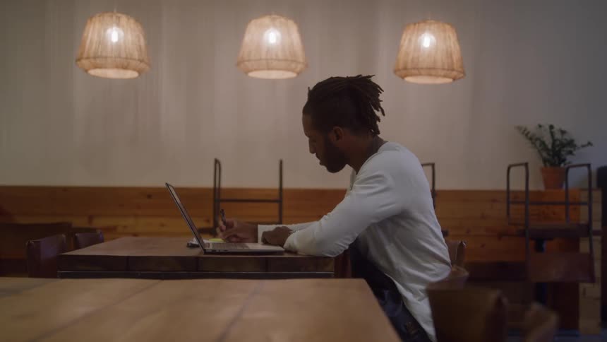 A man with dark hair in dreadlocks sits alone at a long wooden table in a restaurant looking down at a laptop computer and writing something on a notepad
