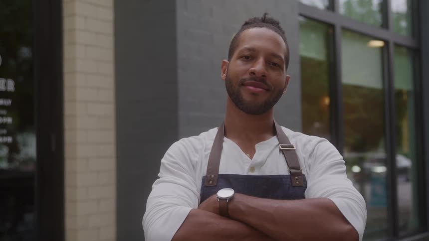A young black male with a beard stands in front of a store at midday wearing an apron as sunlight highlights the storefront behind him