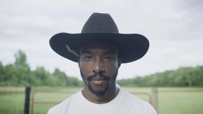 The video is a mediumcloseup shot of a man outdoors wearing a dark cowboy hat and a white tshirt he is looking intently off to the side with a natural outdoor setting visible in the background