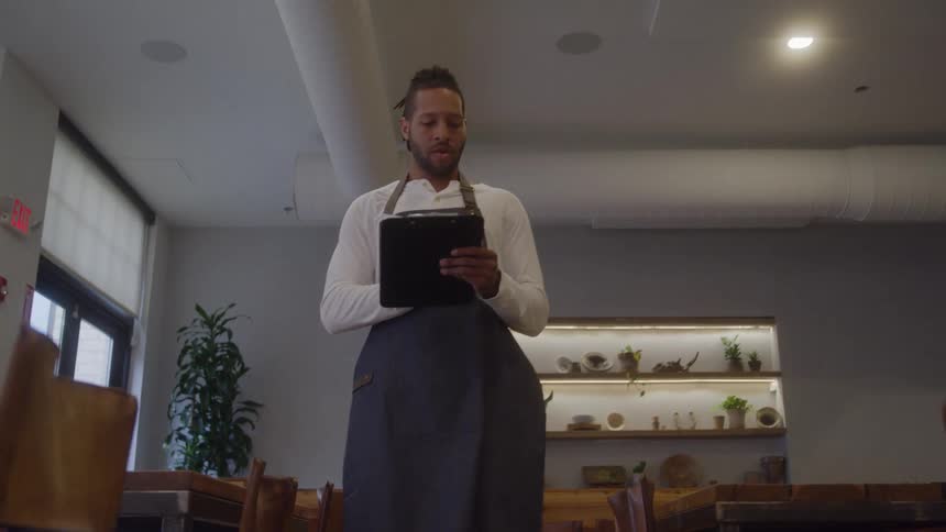 The video is a medium shot of an african american man standing in a restaurant or cafe wearing a white buttondown shirt and a dark apron he is holding a clipboard and gesturing with his hand as if he is taking an order or giving instructions