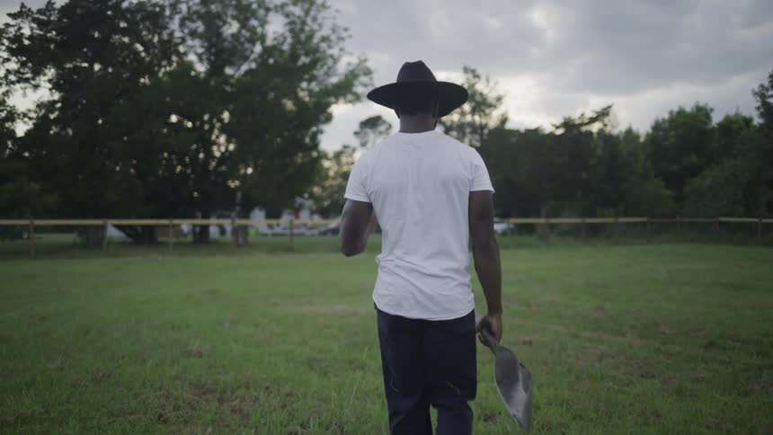 The video shows an african american man walking away from the camera in a grassy field on an overcast day he is wearing a white tshirt dark pants and a dark cowboy hat and he carries a small shovel or trowel in his hand
