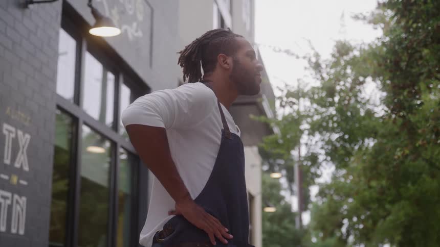 A man with dreadlocks wearing a white shirt and a dark apron stands on a sidewalk next to a brick building storefront with large windows resting his hand on his hip toward green trees under a bright sky