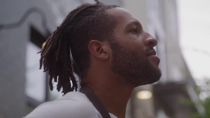The video is a mediumclose up of an african american mans face in profile standing outdoors he has a beard and dreadlocks tied back and he is looking up and slightly away from the camera