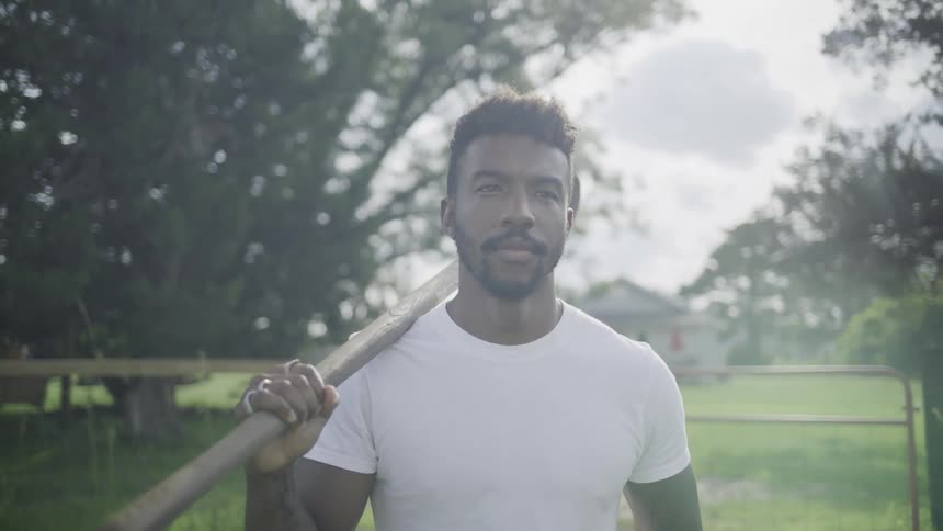 A man wearing a white shortsleeved shirt walks toward the camera on a grassy outdoor path carrying a longhandled shovel resting across his shoulder with green trees and a wooden fence structure visible