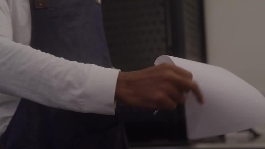 The video is a mediumcloseup shot of a mans torso wearing a white longsleeved shirt and a dark apron as he writes on a clipboard the scene suggests a professional or service environment like a cafe or restaurant where he might be taki