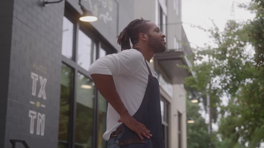 The video is a medium shot of an african american man in a white longsleeve shirt and a dark blue apron standing outside a business storefront he is looking up and to the side with a smile appearing relaxed or taking a break