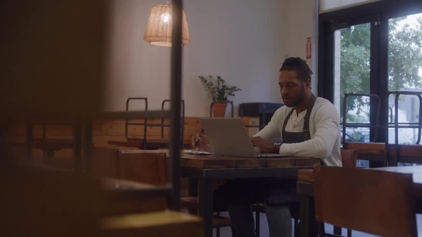 The video is a medium shot of an african american man working at a wooden table with a laptop and a notebook in what looks like a welllit cafe or restaurant interior he has his hair tied back and is wearing a white longsleeved shirt with a dark
