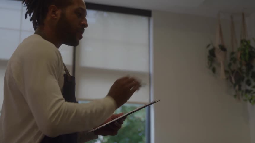 The video is a mediumcloseup of an african american man working inside a welllit space wearing a white longsleeve shirt and a dark apron he is standing in front of a window writing notes on a clipboard