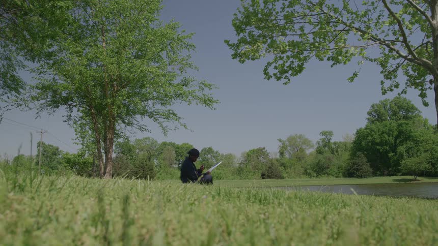 The video is a wide shot of an african american man sitting on the grass outdoors on a sunny day he is wearing dark clothing and a cap and he is painting on a white canvas or drawing board by the edge of a pond or small lake