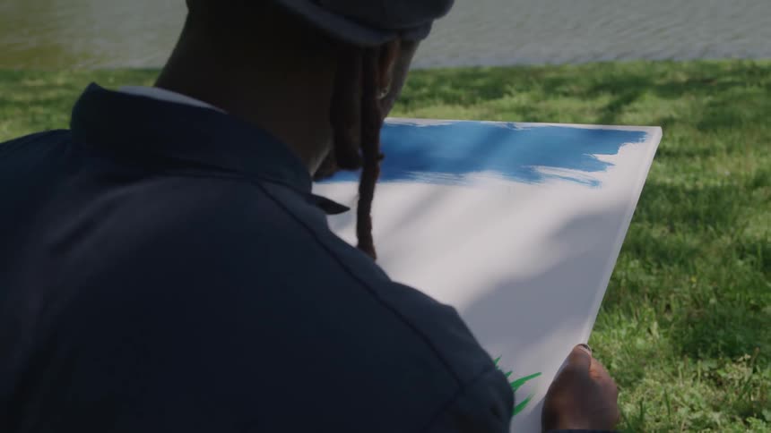 The video is a closeup of an african american man sitting on the grass painting on a white canvas with blue and green paint he is shown in profile and from behind wearing a dark jacket and cap with his dreadlocks visible as he leans over the c