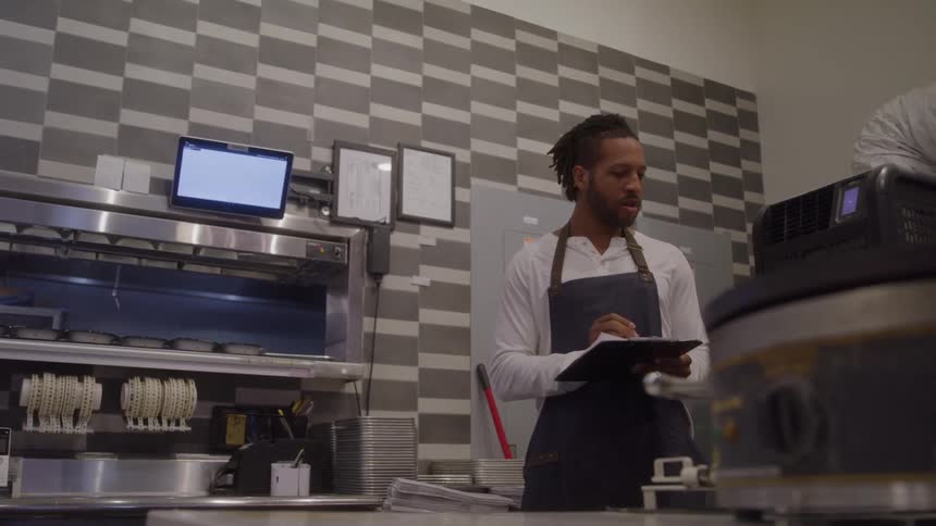 A man wearing a white longsleeved shirt and a dark apron stands behind a metal counter in a commercial kitchen holding a clipboard and reading and writing on the paper with cooking equipment and a patterned tile wall visible in the background