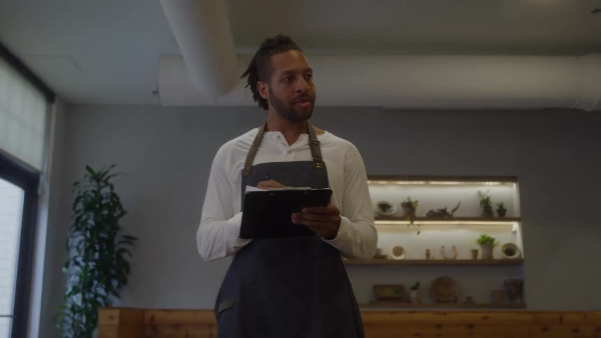 A man wearing a white longsleeved shirt and a dark apron stands in an indoor commercial space holding a clipboard and writing on it while looking up and around the space which has wooden paneling and illuminated shelves