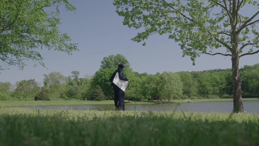 A man wearing dark clothing walks from the left side of the frame into the center of a grassy field carrying a large canvas under his arm then stops facing the water and bends over to place the object on the ground