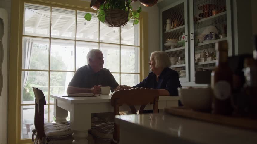 The video is a stationary shot of an elderly couple seated at a bright kitchen table drinking coffee or tea they are facing each other and appear to be conversing pleasantly in a warm sunlit room with large windows