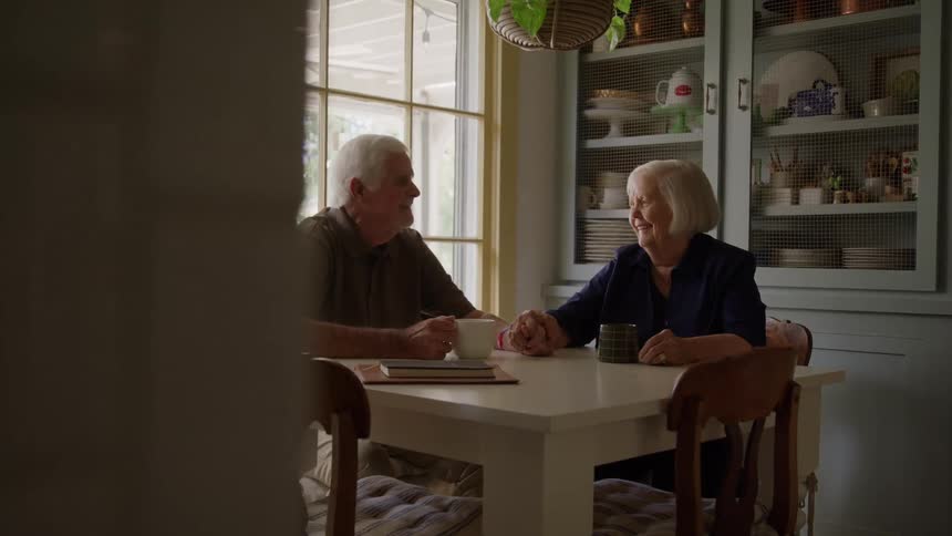 An elderly couple sit facing each other holding hands across the table and smiling with a china cabinet filled with dishes visible in the background and bright daylight streaming from the window