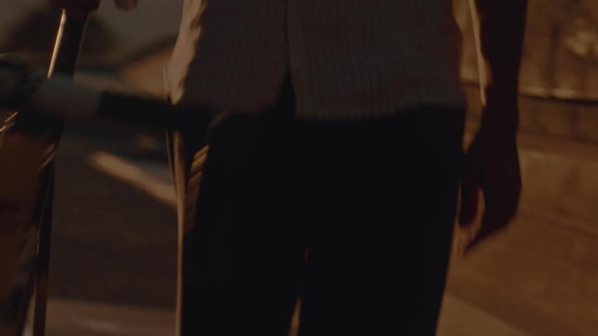 A person is pushing a darkcolored road bicycle forward in a tight lowlight shot with the camera focusing on the back half of the bike and the persons lower body in a urban setting